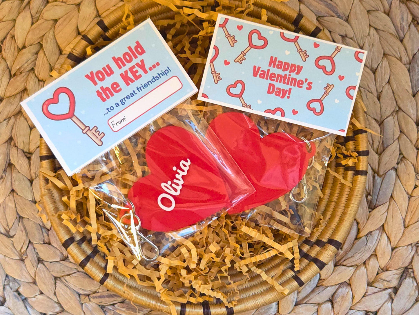 Two classroom valentines are pictured in a dish of kraft paper on a woven background. Each is a personalized heart keychain, which could also be used as a zipper pull or bag charm. It features the child's name, in this case the name Olivia. At the top of the bag, the bag topper serves as the Valentine with the message "You hold the key to a great friendship" with space for the child to write who the gift is from. On the back it says "Happy Valentine's Day"