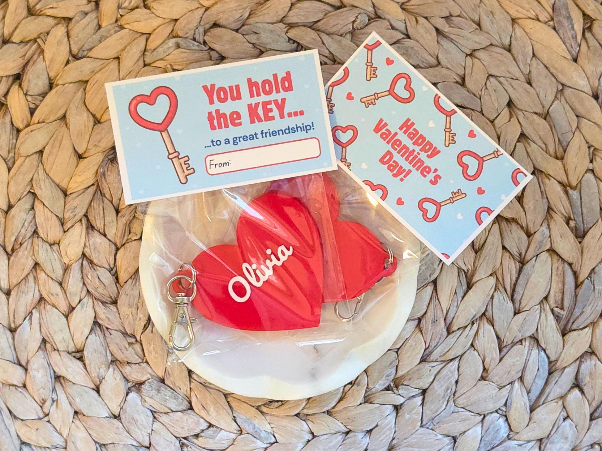 Two classroom valentines are pictured in a white dish on a woven background. Each is a personalized heart keychain, which could also be used as a zipper pull or bag charm. It features the child's name, in this case the name Olivia. At the top of the bag, the bag topper serves as the Valentine with the message "You hold the key to a great friendship" with space for the child to write who the gift is from. On the back it says "Happy Valentine's Day"