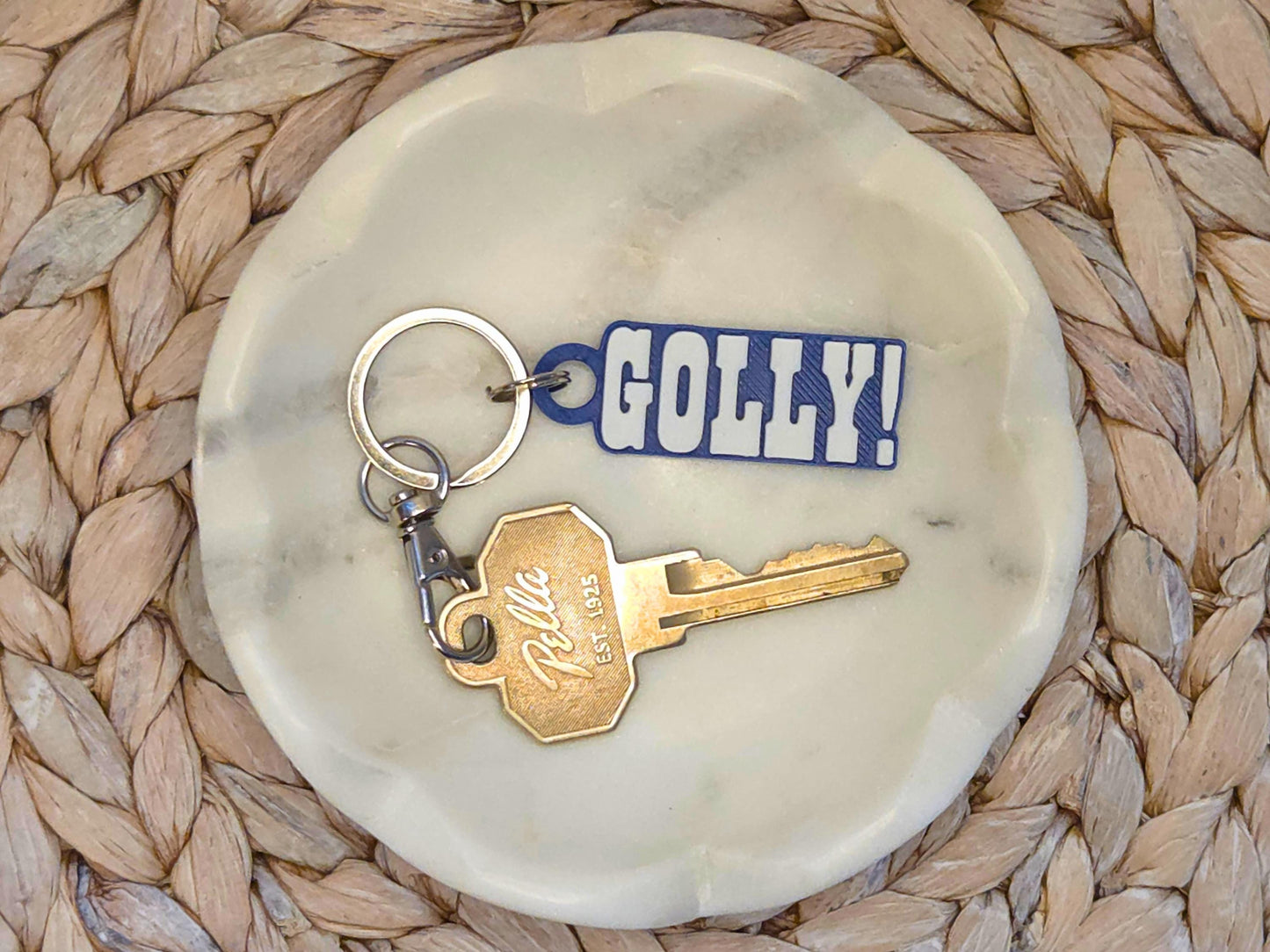 Indianpolis Quarterback Keychain featuring mic'd up language Golly! in white text on a blue background. Pictured in a white dish with a key for scale, on a woven background.