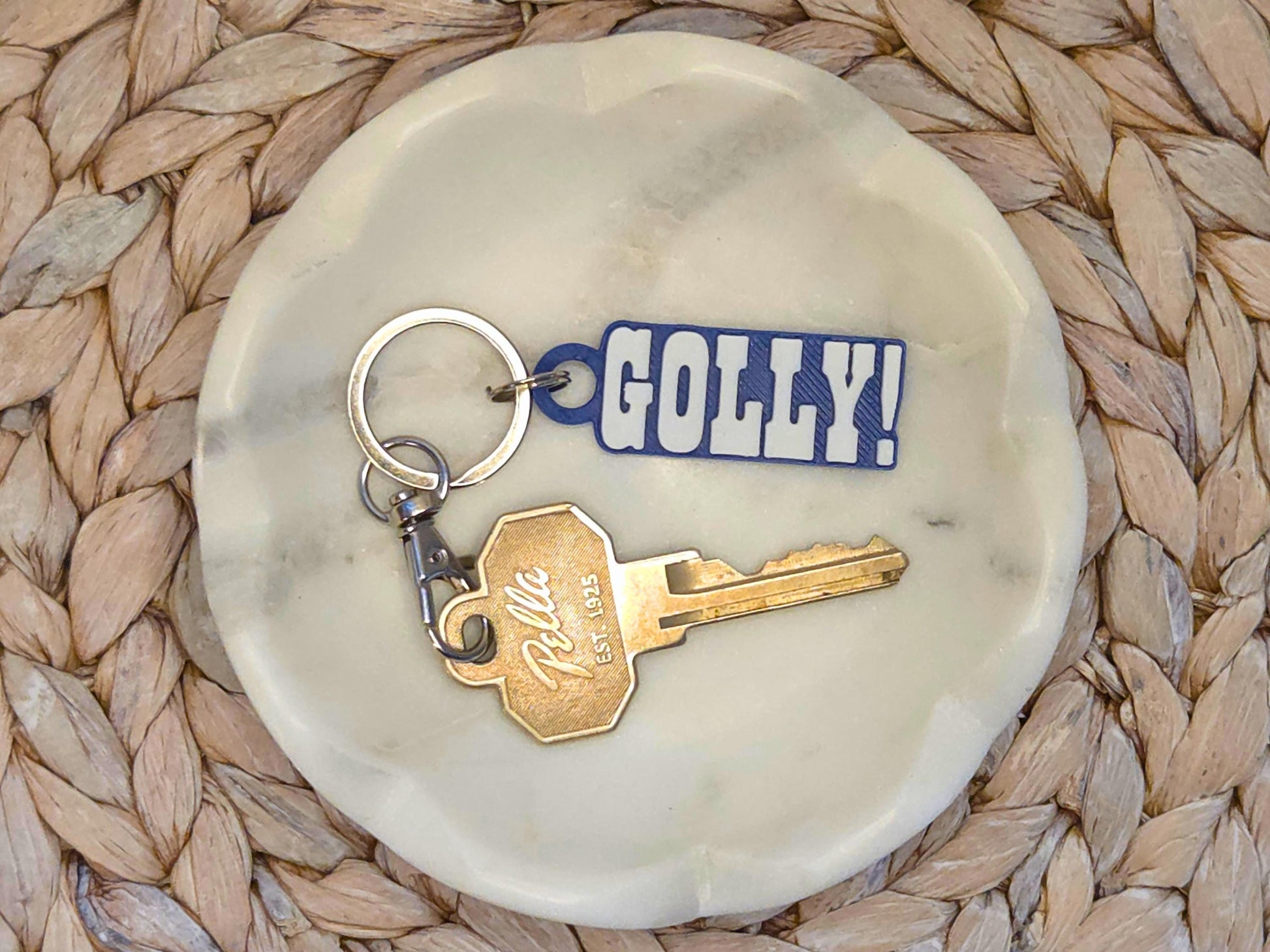 Indianpolis Quarterback Keychain featuring mic'd up language Golly! in white text on a blue background. Pictured in a white dish with a key for scale, on a woven background.