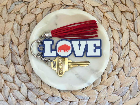 Buffalo Love Keychain or bag tag is shown in a white dish on a woven background with optional red tassel. A key is shown for scale.