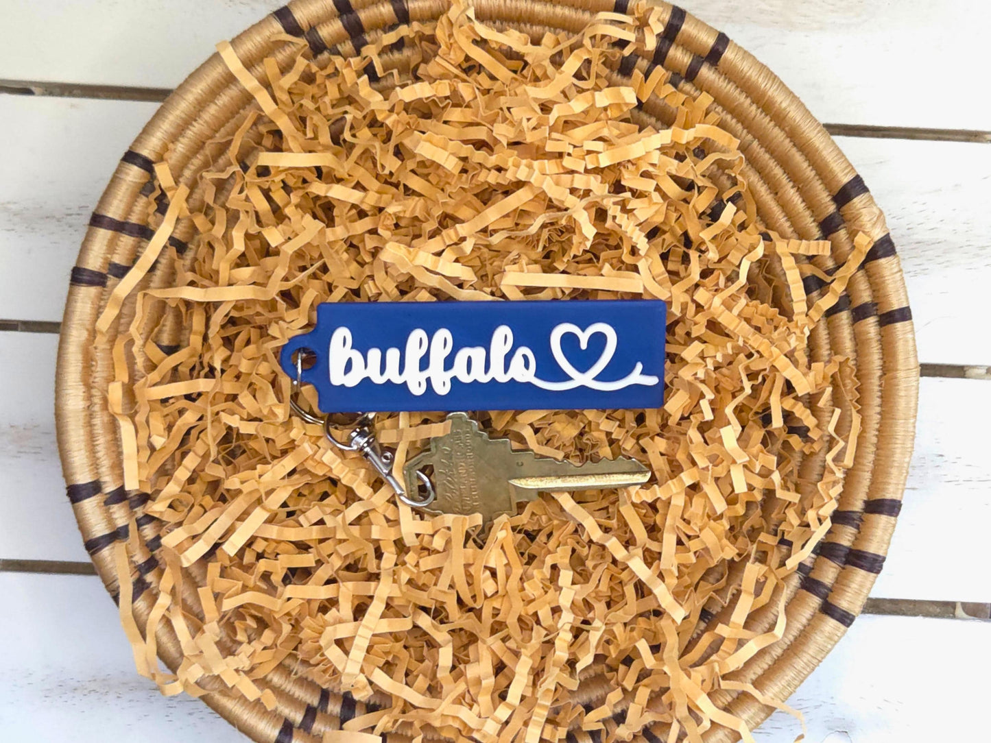 Buffalo heart keychain is featured in a basket with kraft paper on a wooden background. A key is shown for scale.