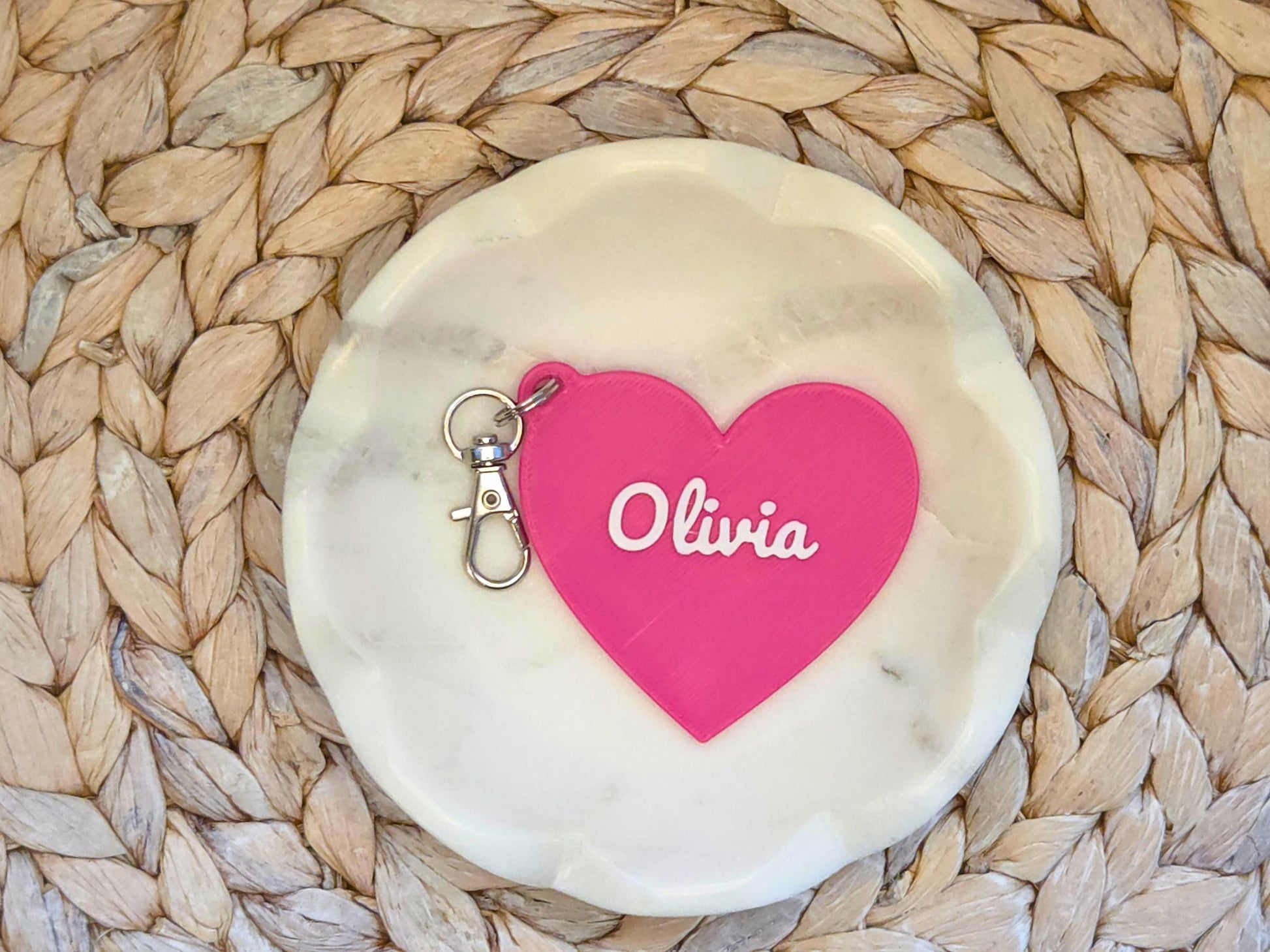 The front of the personalized heart keychain in pink is pictured in a white dish on a woven background. It can also be used as a zipper pull or bag charm. It features the child's name, in this case the name Olivia in script font.