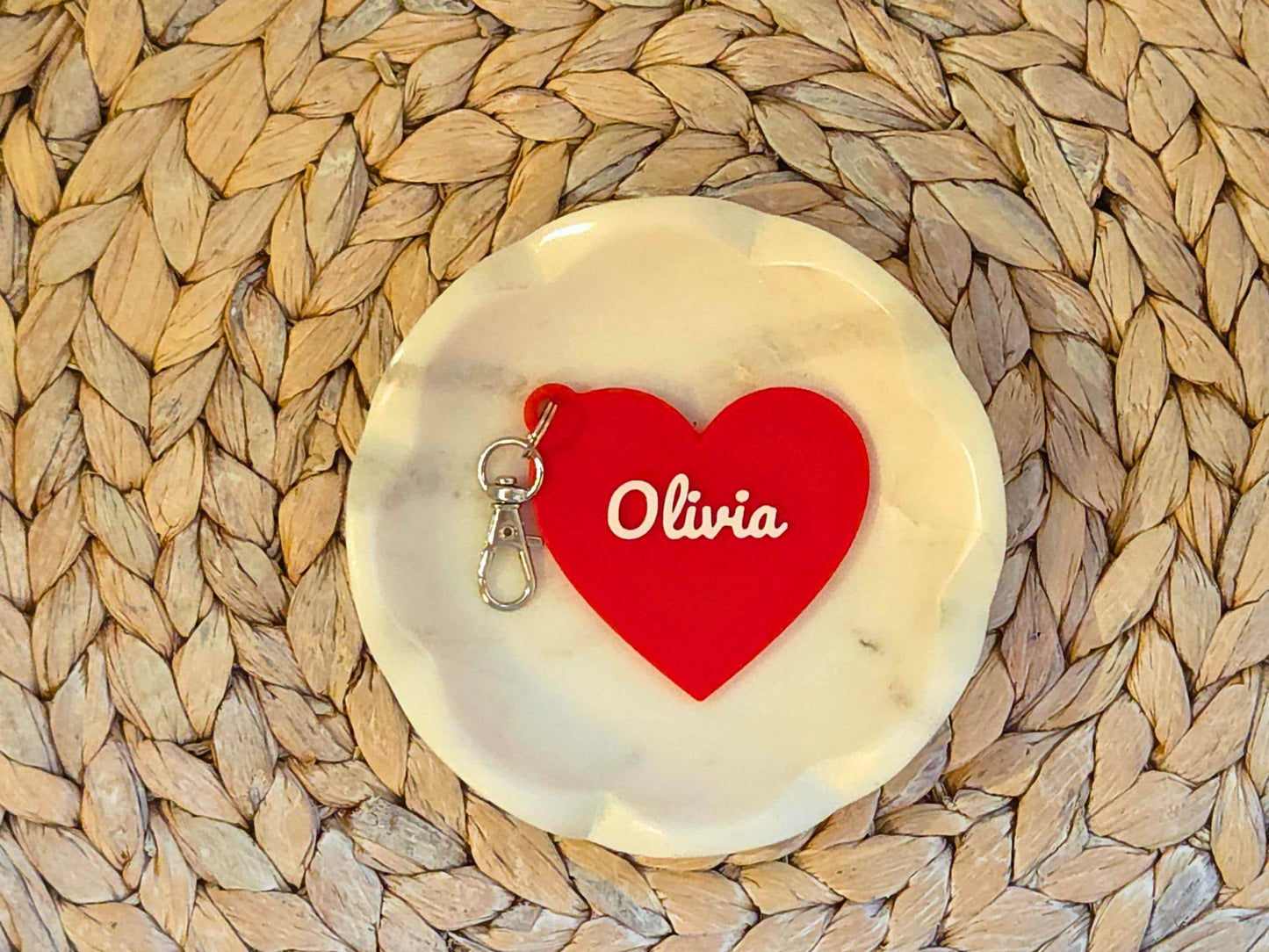 The front of the personalized heart keychain in red is pictured in a white dish on a woven background. It can also be used as a zipper pull or bag charm. It features the child's name, in this case the name Olivia in script font.
