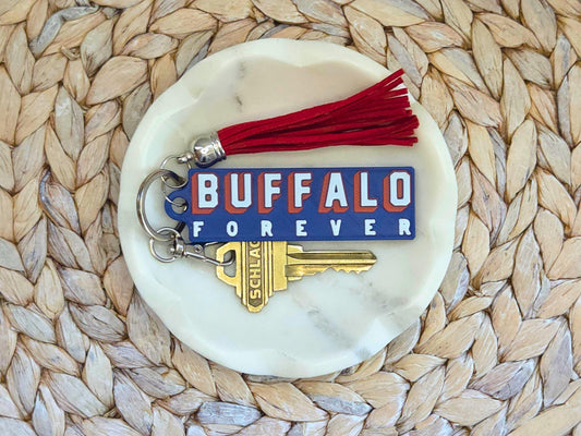 Buffalo Forever keychain is displayed in a white dish on a woven background with an optional red tassel. A key is shown for size.