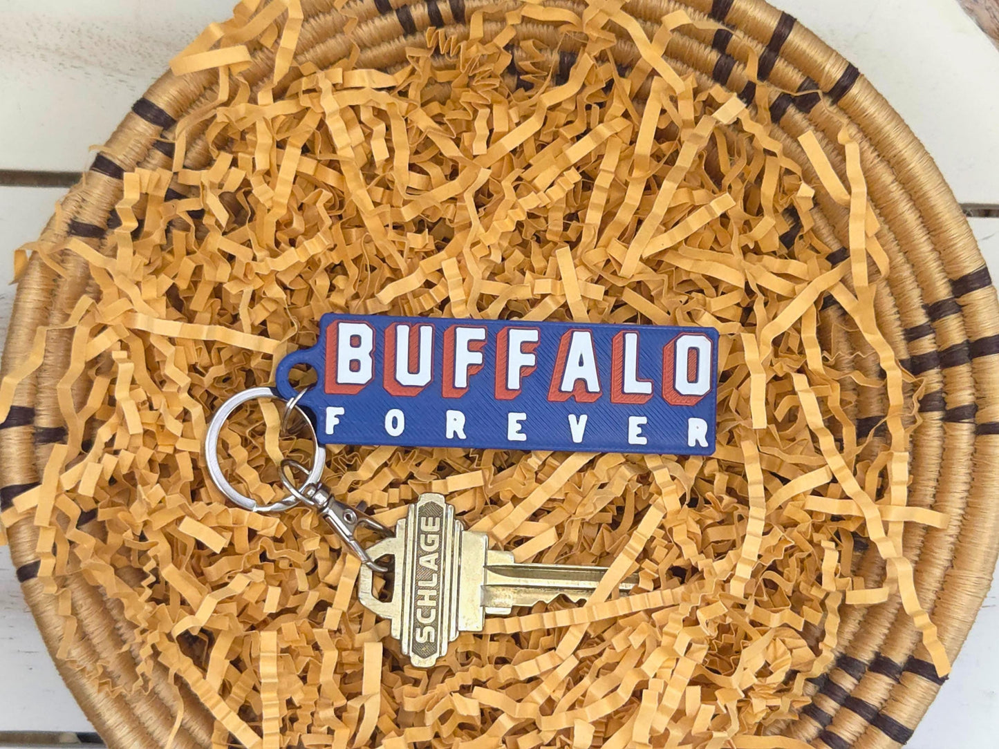 Buffalo Forever keychain is displayed in a basket with kraft paper on a wooden background. A key is shown for size.