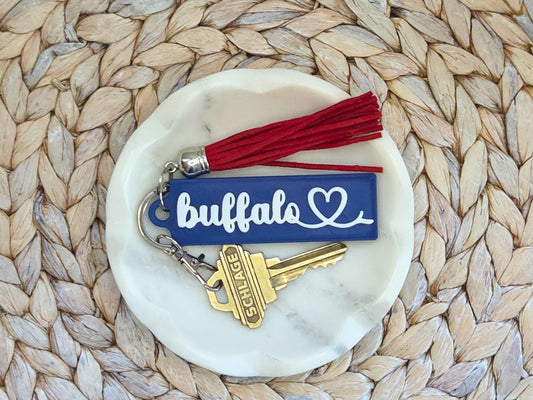 Buffalo heart keychain is featured in a white dish on a woven background with optional red tassel. A key is shown for scale.