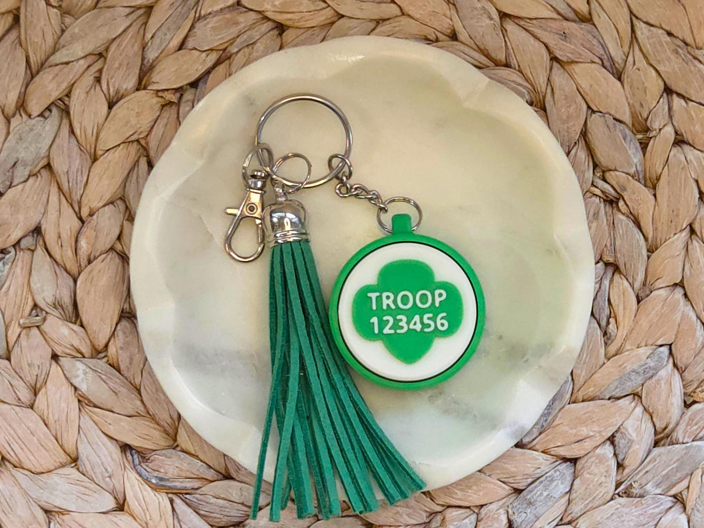 Green and white scout fidget clicker keychain personalized with the troop number 12345 and featured with an optional green tassel is shown in a white dish on a woven background.