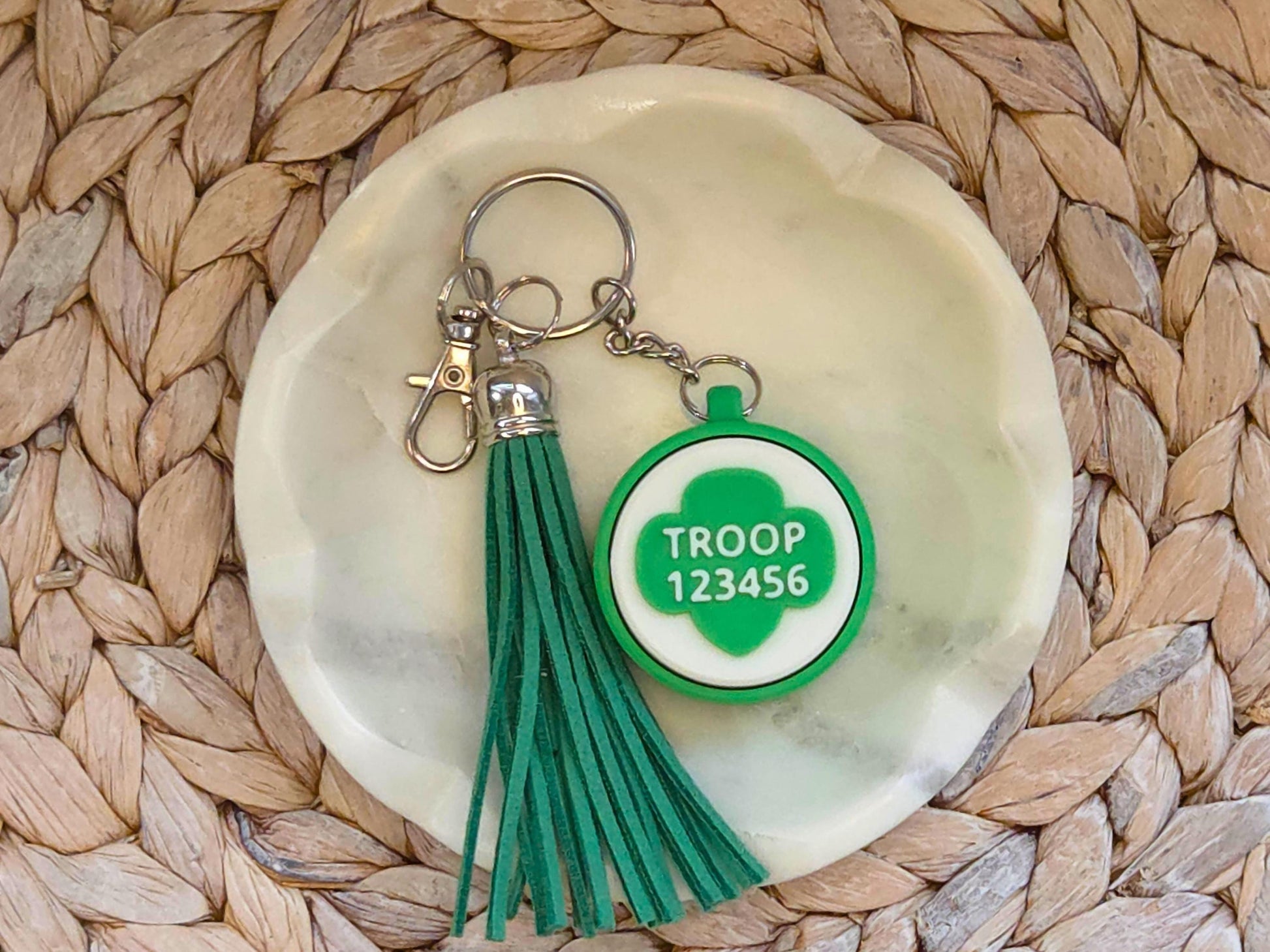 Green and white scout fidget clicker keychain personalized with the troop number 12345 and featured with an optional green tassel is shown in a white dish on a woven background.