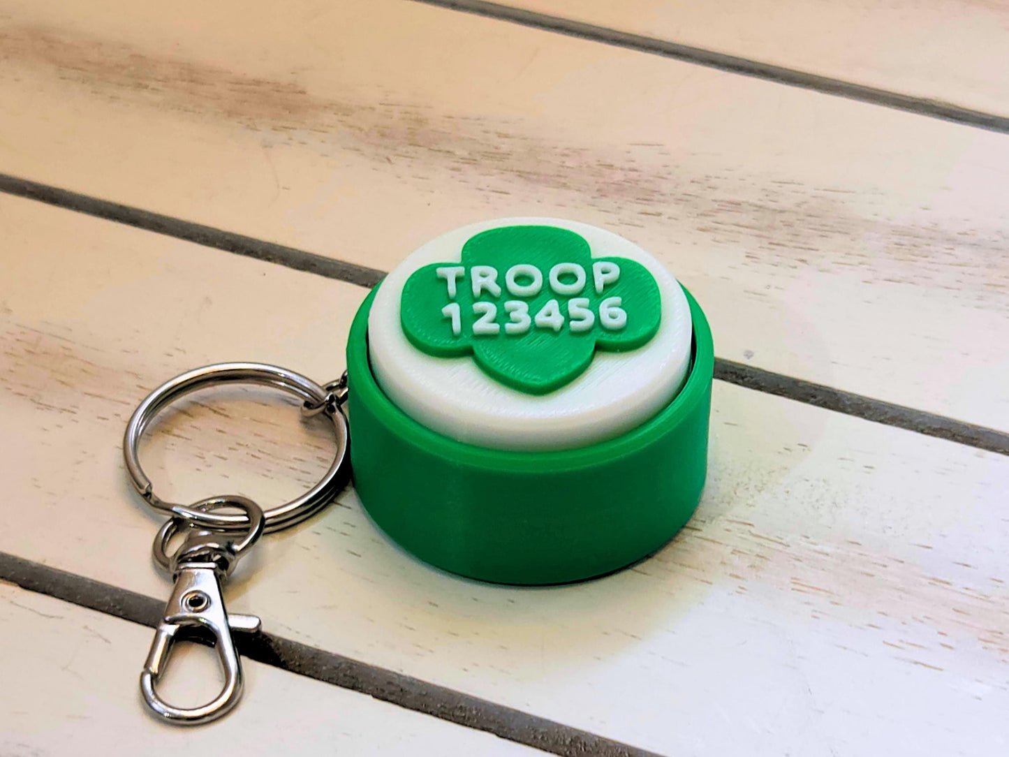 A green and white scout fidget clicker keychain is shown on a wooden background. It is personalized with troop number 12345 in raised white lettering.