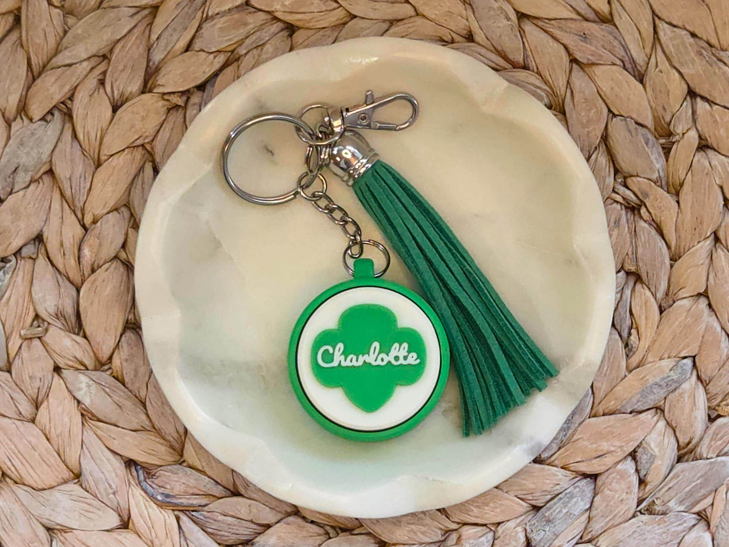 Green and white scout fidget clicker keychain personalized with the name Charlotte and featured with an optional green tassel is shown in a white dish on a woven background.