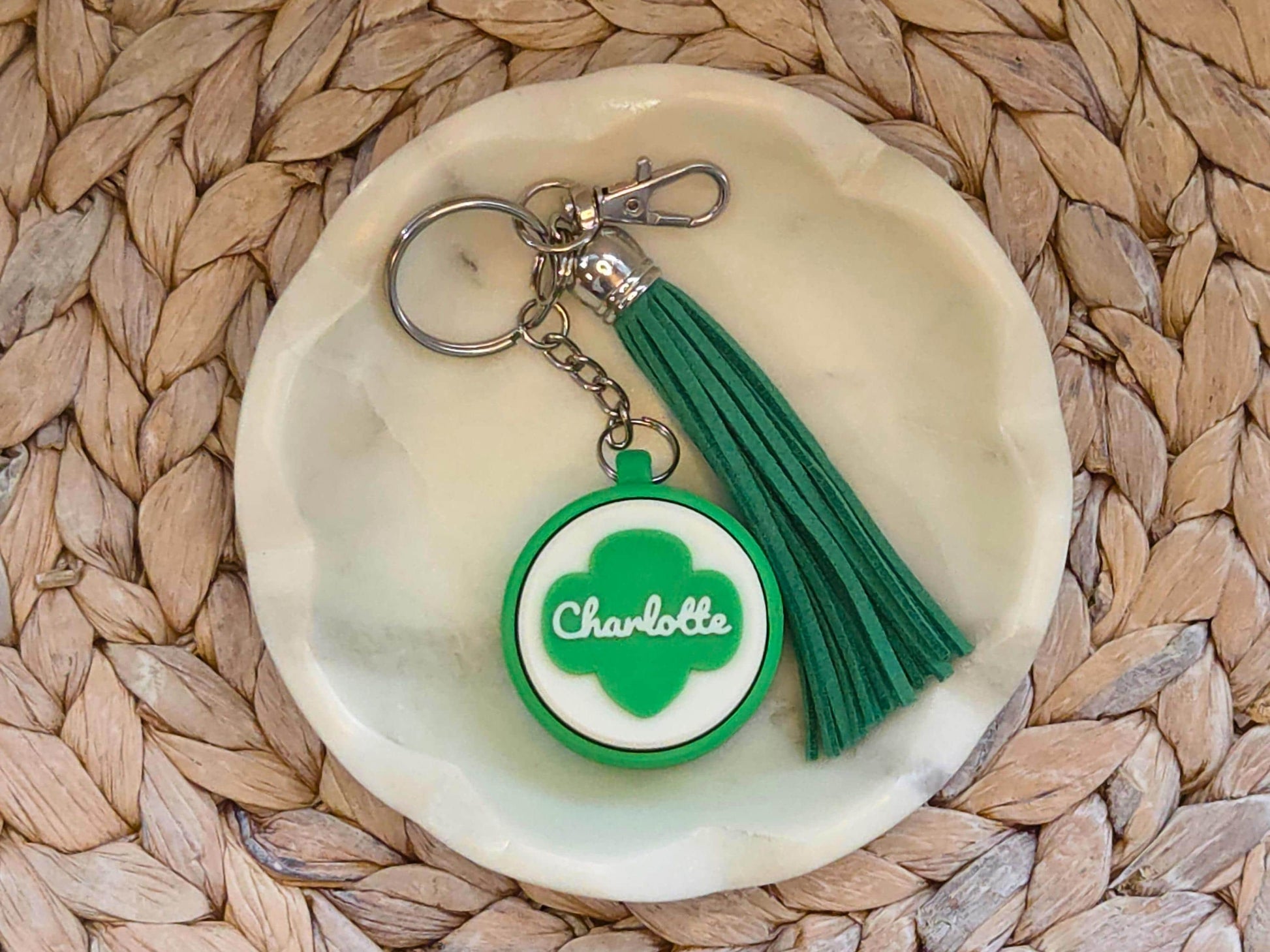 Green and white scout fidget clicker keychain personalized with the name Charlotte and featured with an optional green tassel is shown in a white dish on a woven background.