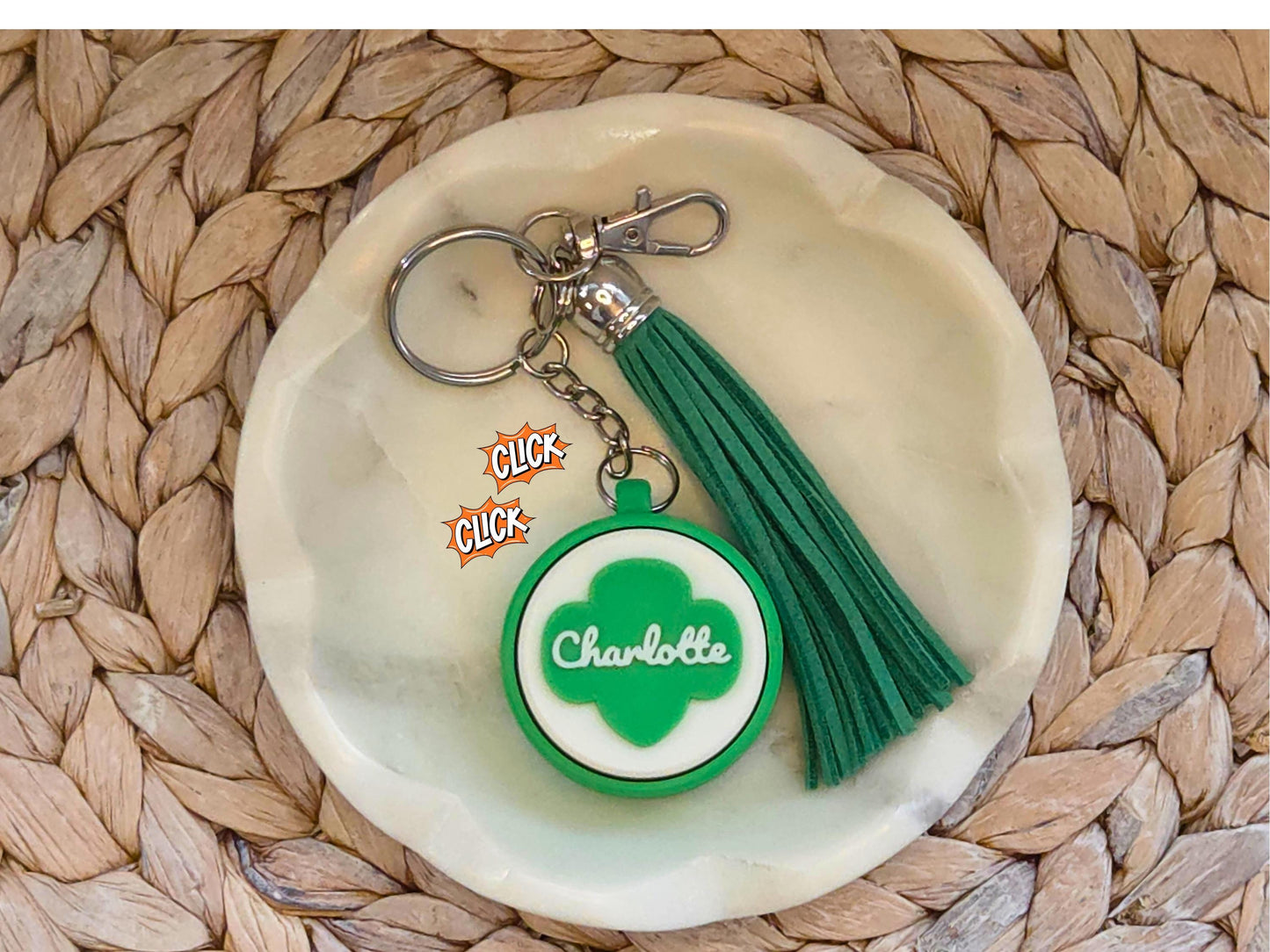 Green and white scout fidget clicker keychain personalized with the name Charlotte and featured with an optional green tassel is shown in a white dish on a woven background.
