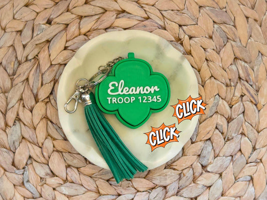 Picture of green girl scout fidget clicker keychain with optional green tassel in a white dish on a woven background. The keychain is personalized with the name Eleanor and the troop number 12345.