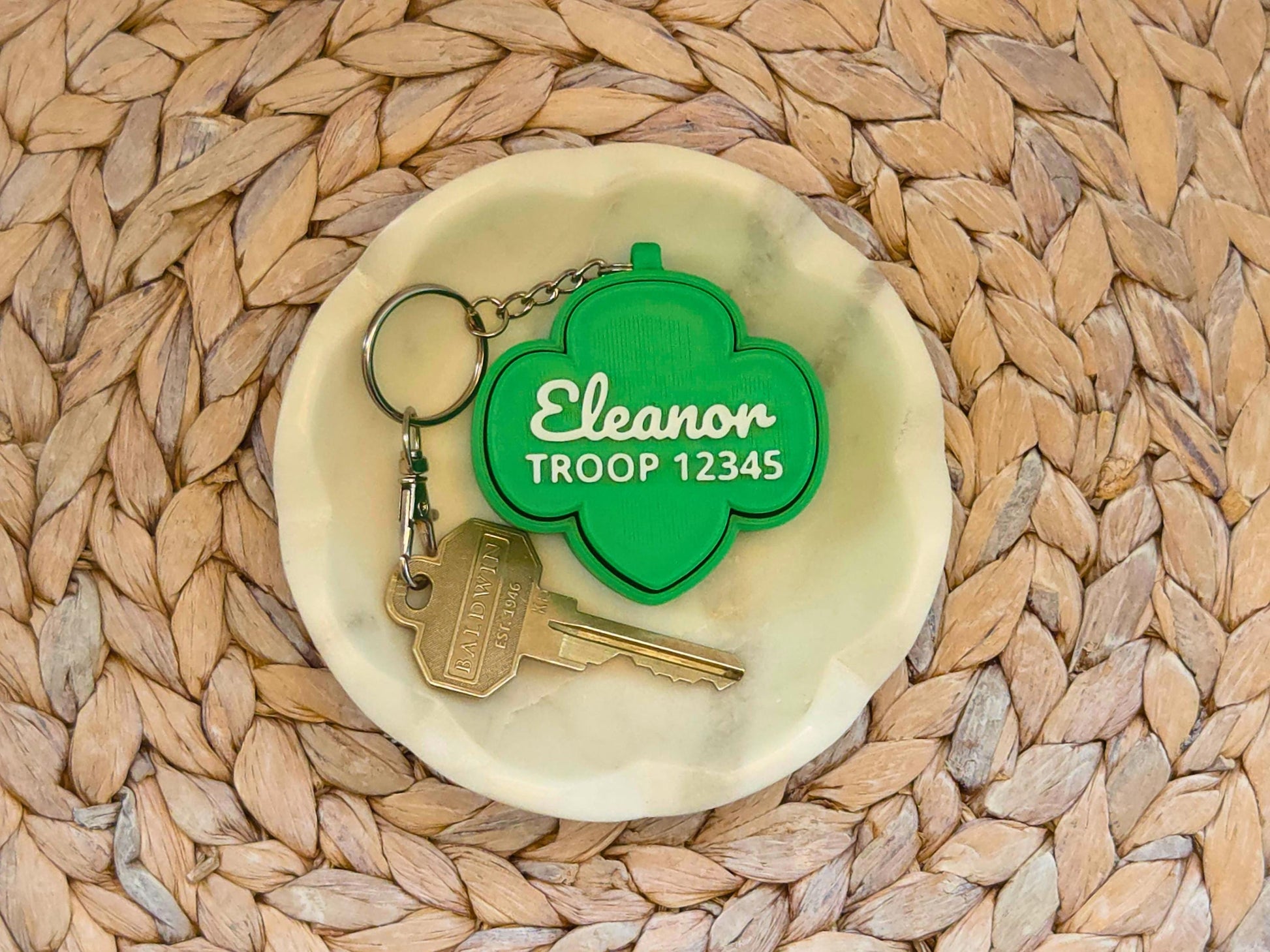 Picture of green girl scout fidget clicker keychain in a white dish on a woven background and a key shown for scale. The keychain is personalized with the name Eleanor and the troop number 12345.