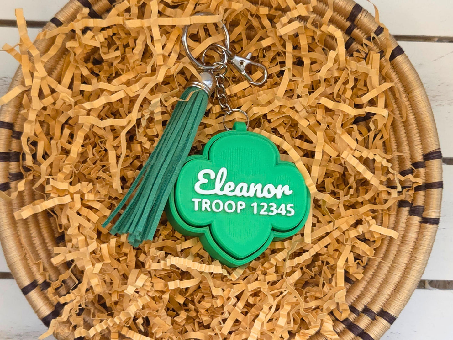 Picture of green girl scout fidget clicker keychain with optional green tassel in basket of kraft paper on a wooden background. The keychain is personalized with the name Eleanor and the troop number 12345.