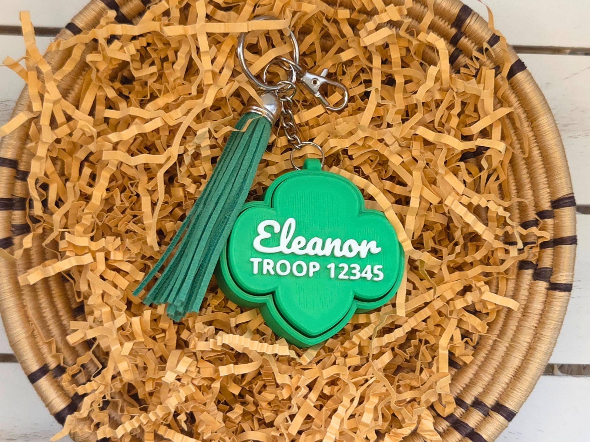 Picture of green girl scout fidget clicker keychain with optional green tassel in basket of kraft paper on a wooden background. The keychain is personalized with the name Eleanor and the troop number 12345.