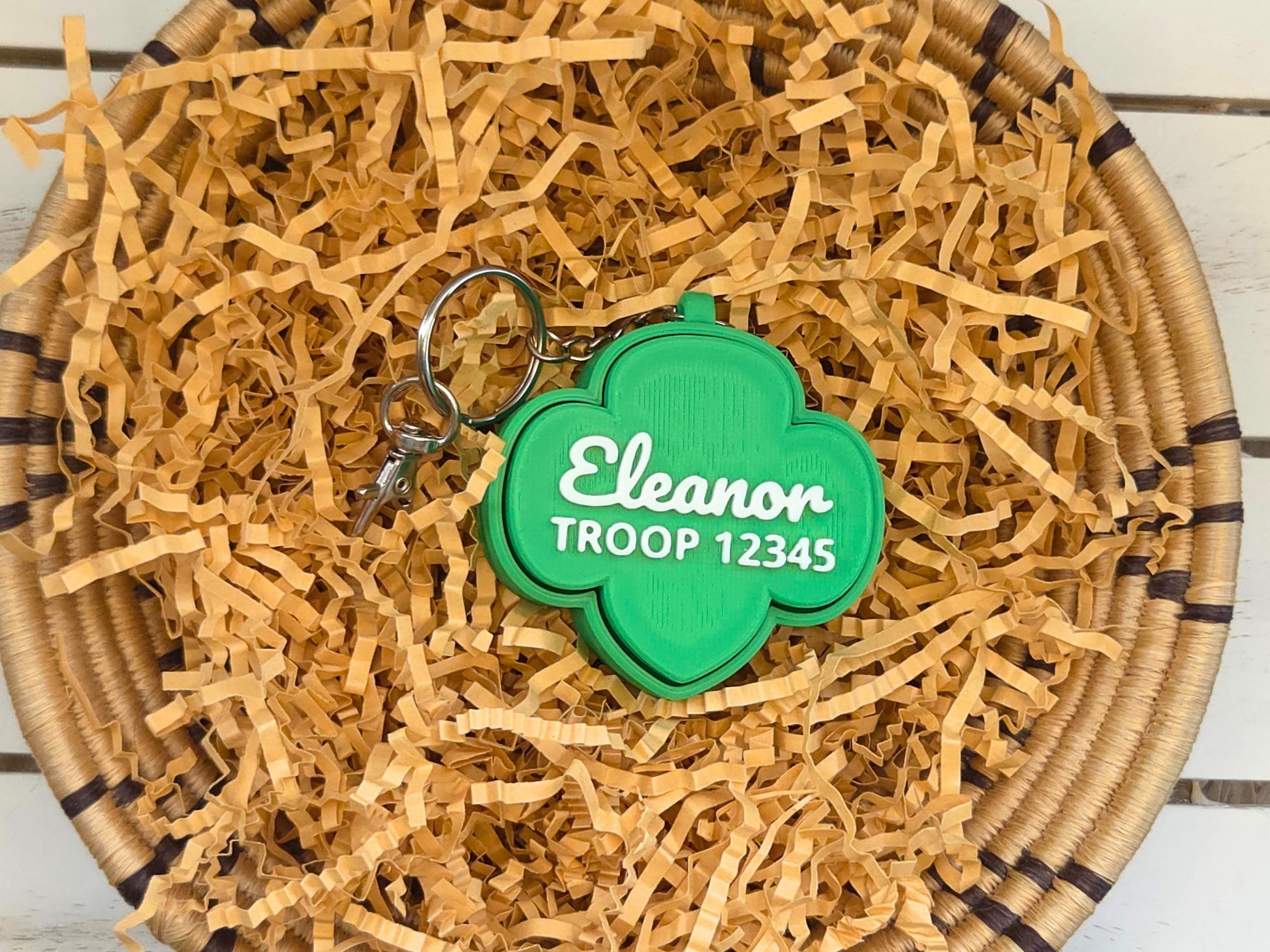 Picture of green girl scout fidget clicker keychain in basket of kraft paper on a wooden background. The keychain is personalized with the name Eleanor and the troop number 12345.