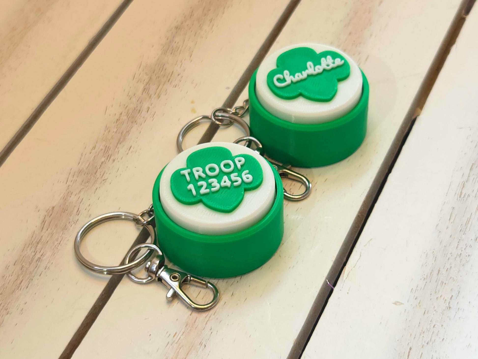Two green and white scout fidget clicker keychains are shown on a white wooden background. One is personalized with the name Charlotte and the other is personalized with troop number 12345.