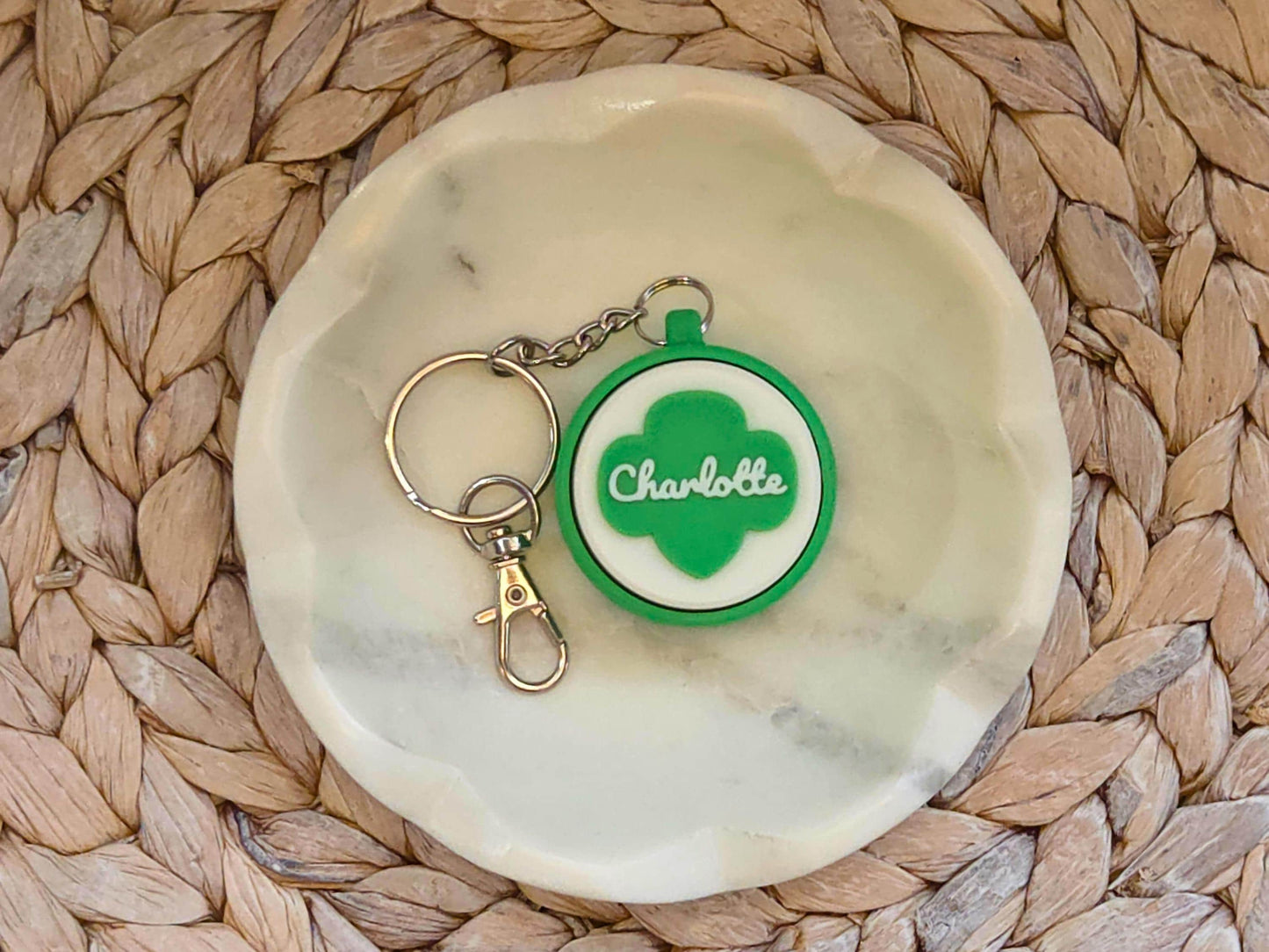 Green and white scout fidget clicker keychain personalized with the name Charlotte is shown in a white dish on a woven background.