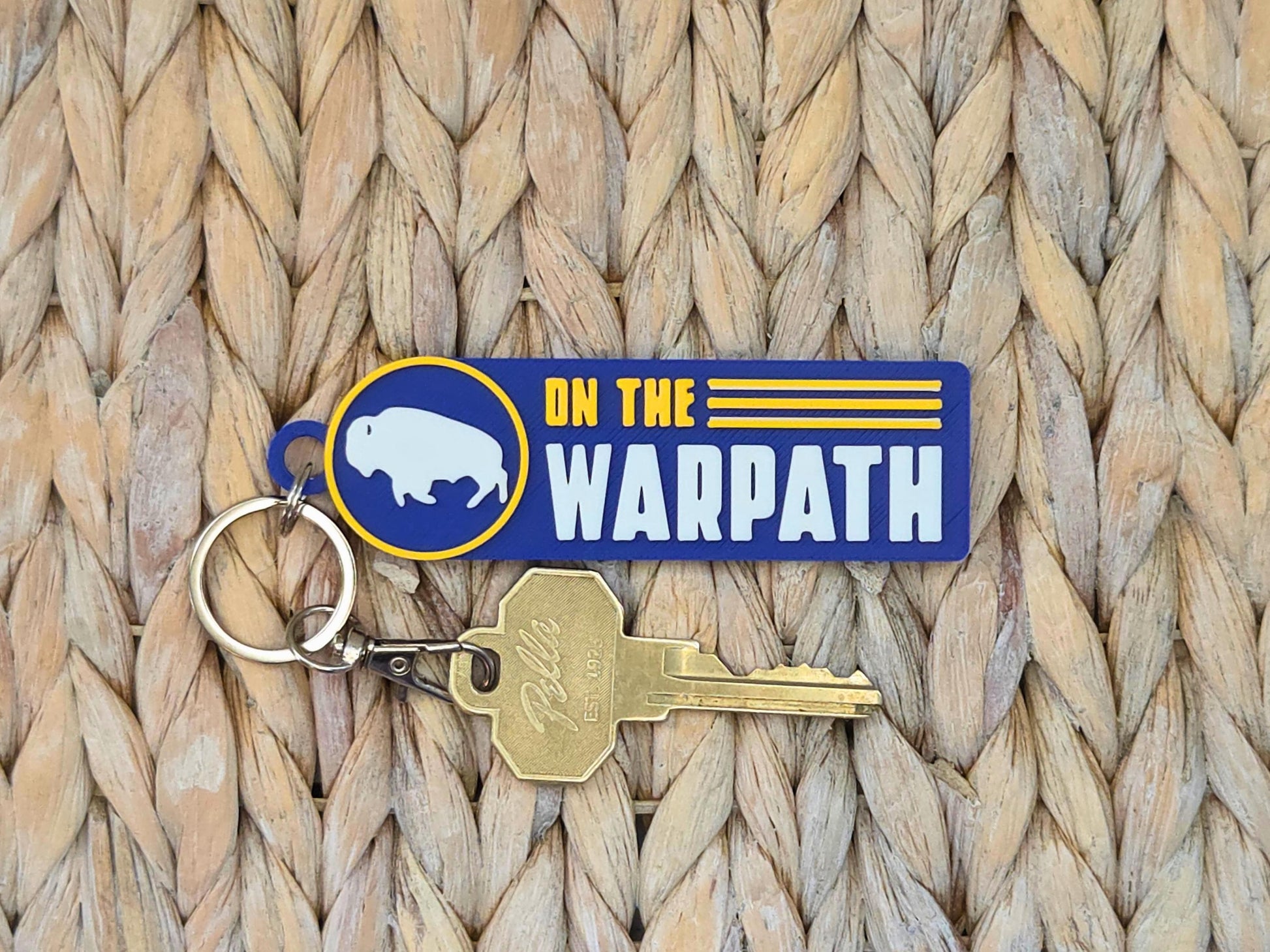 Close-up of durable "On the Warpath" 3D-printed bag tag in blue and gold on a natural woven straw surface.