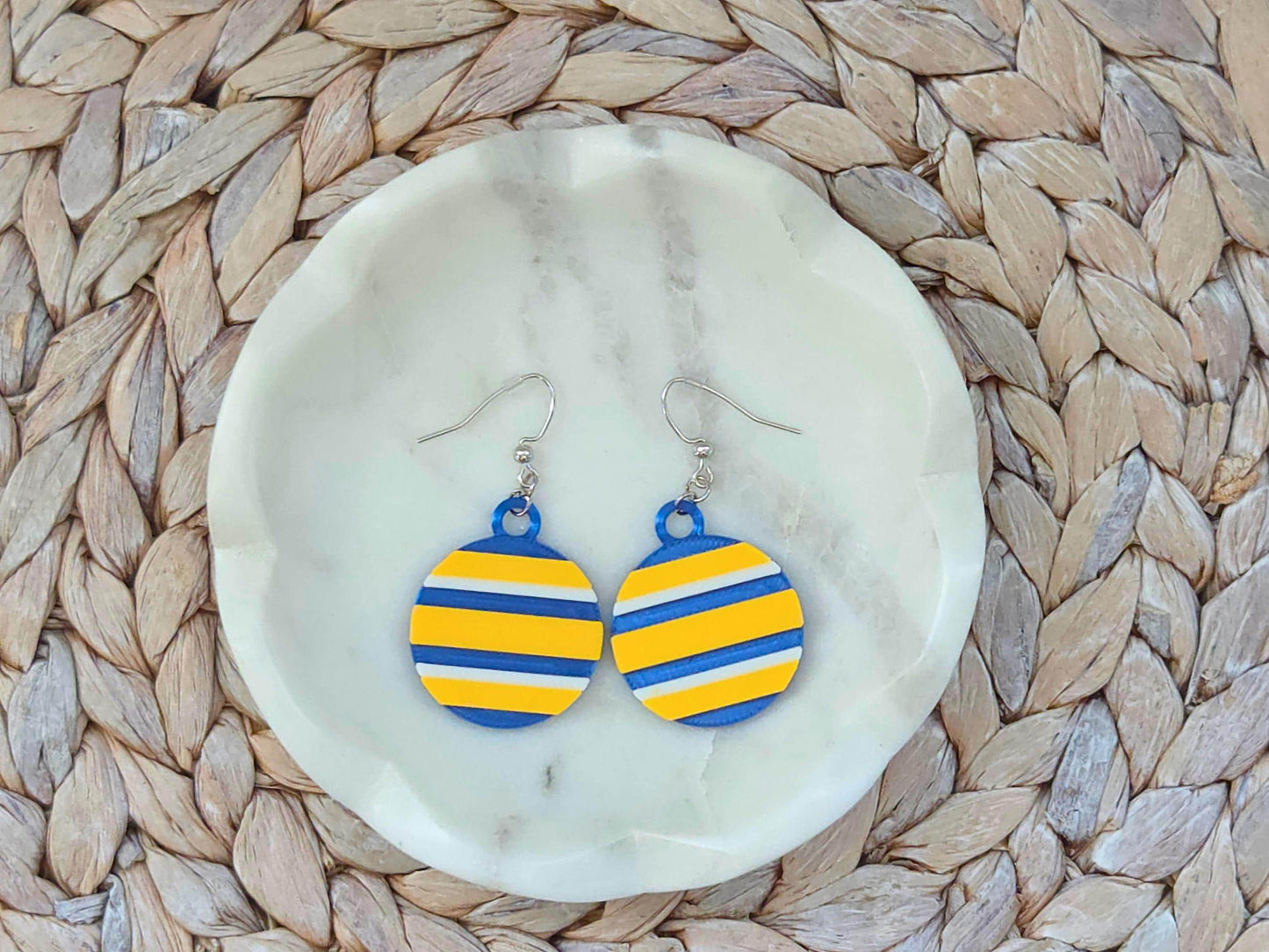 Pair of circular Buffalo hockey earrings with blue, yellow, and white stripes on a white marble jewelry dish.