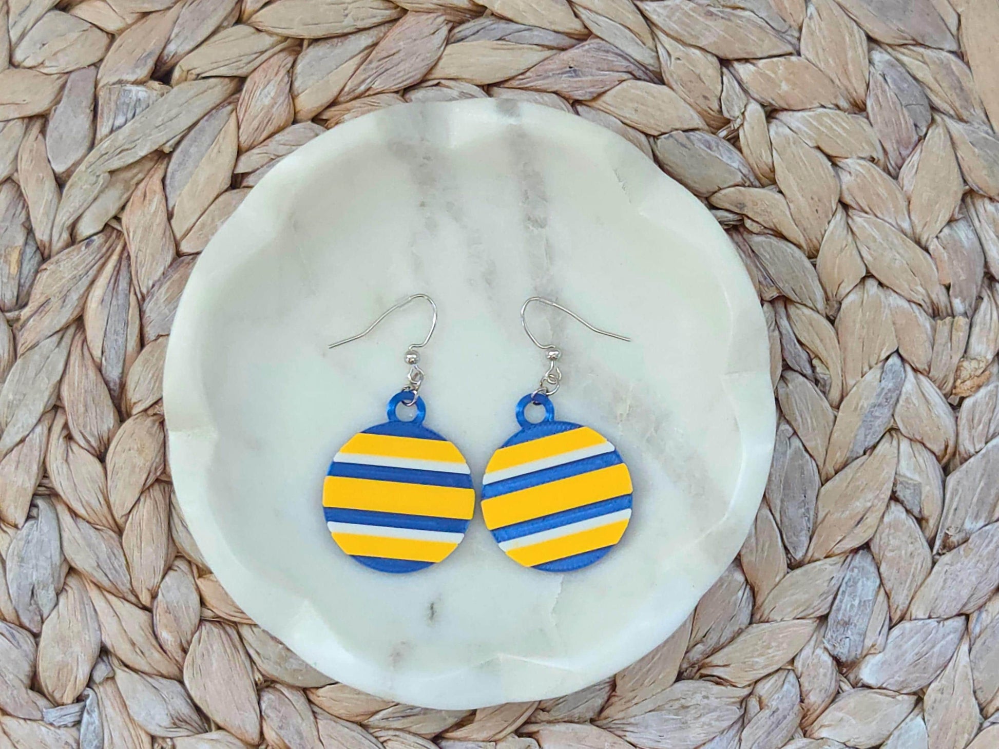 Pair of circular Buffalo hockey earrings with blue, yellow, and white stripes on a white marble jewelry dish.
