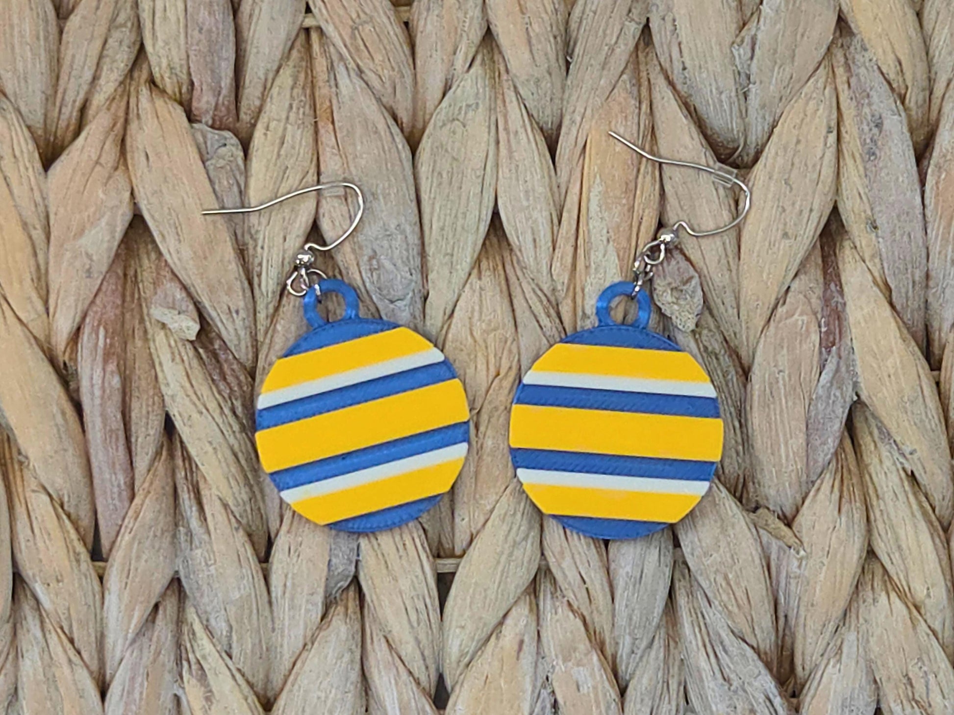 Close-up of 3D-printed striped disc earrings in blue and gold team colors on a natural woven straw background.
