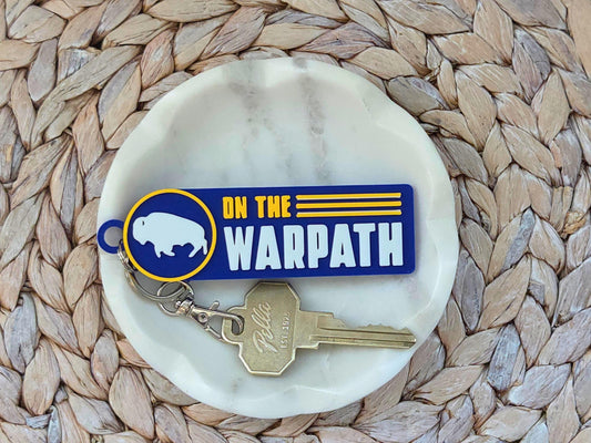 Blue and yellow 3D-printed keychain with "ON THE WARPATH" text and white bison logo on a marble jewelry dish.