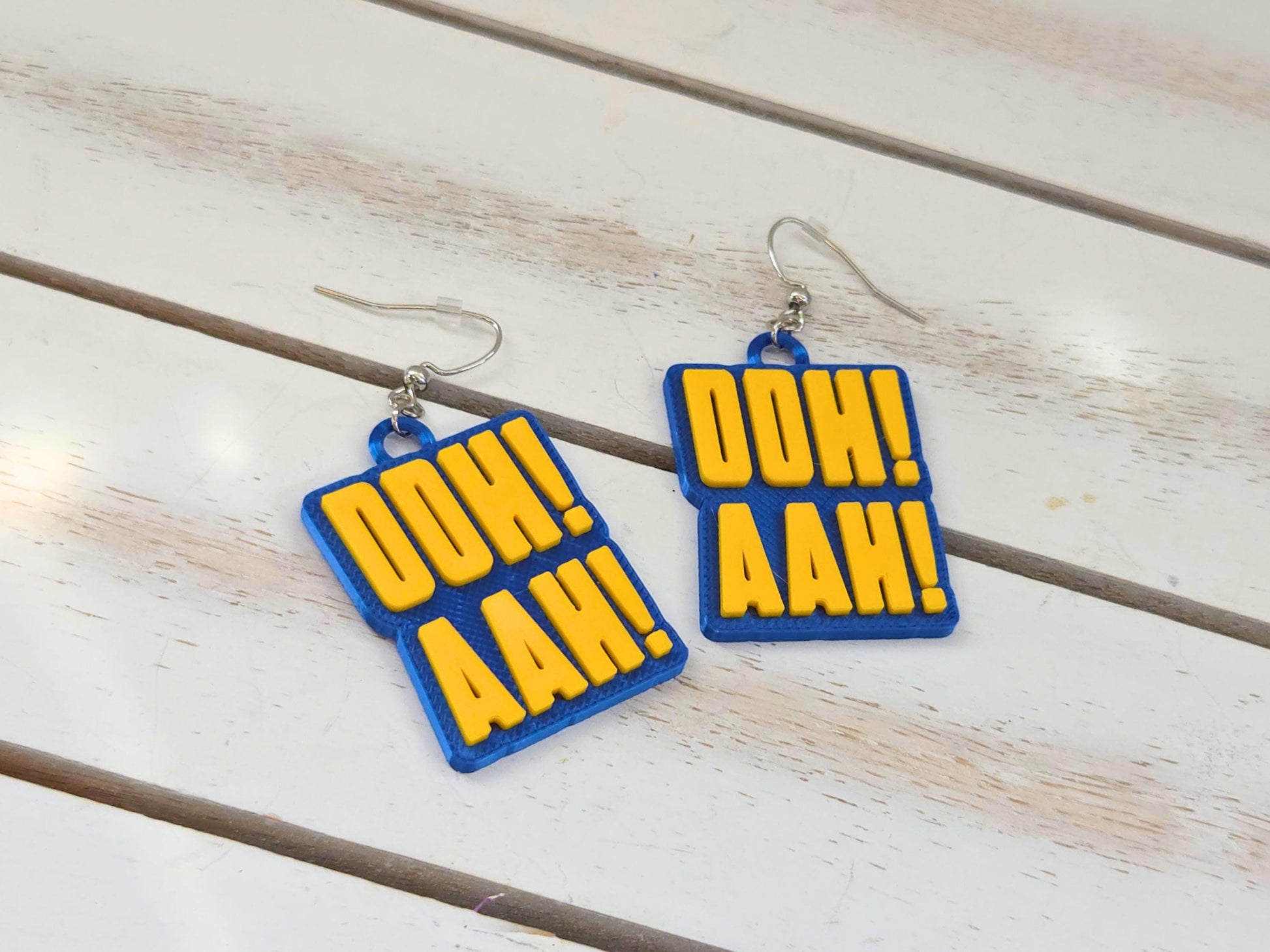 3D-printed Buffalo hockey earrings featuring yellow "OOH! AAH!" text on a blue rectangular base, displayed on white wood.