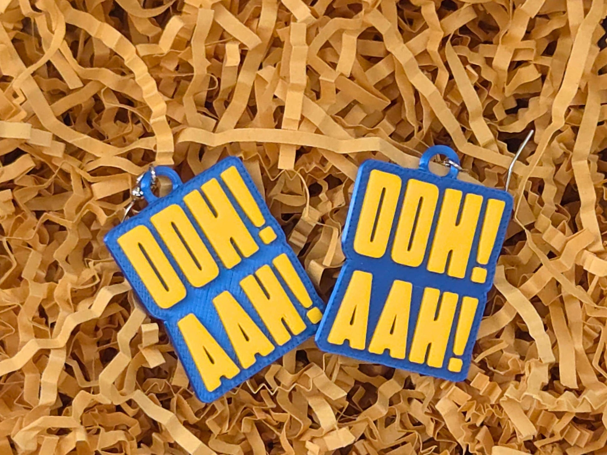 Blue and yellow "OOH! AAH!" fan earrings resting on tan gift basket filler, ideal for hockey-themed stocking stuffers.