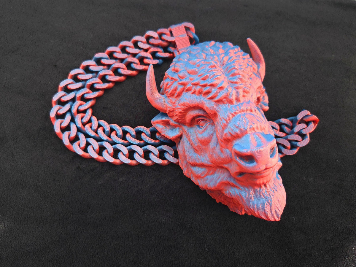 A red and blue 3D printed buffalo head necklace with a thick link chain laid out flat on a black surface.