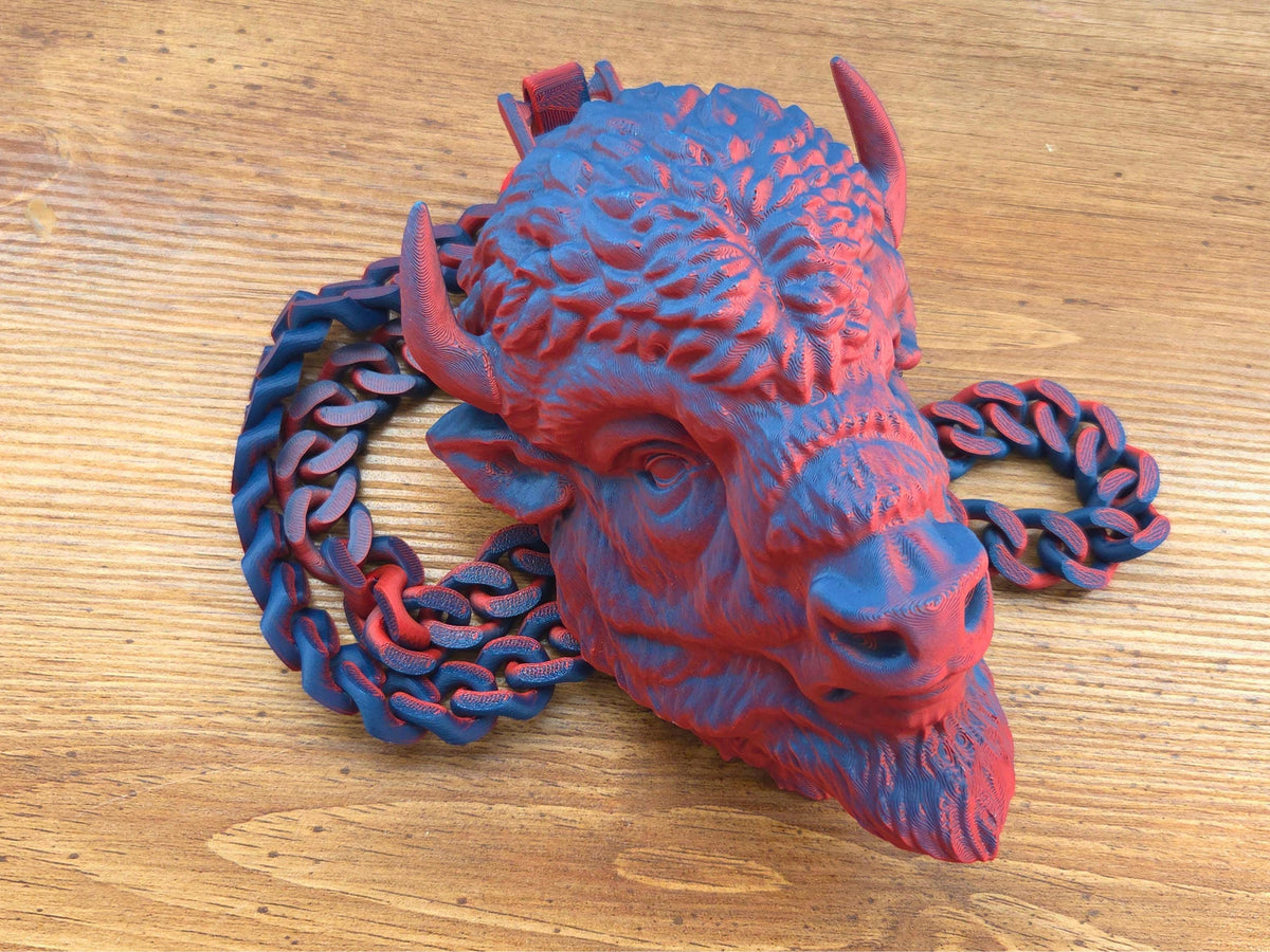 A 3D printed red and blue buffalo head pendant attached to a thick matching link chain, resting on a wooden surface.